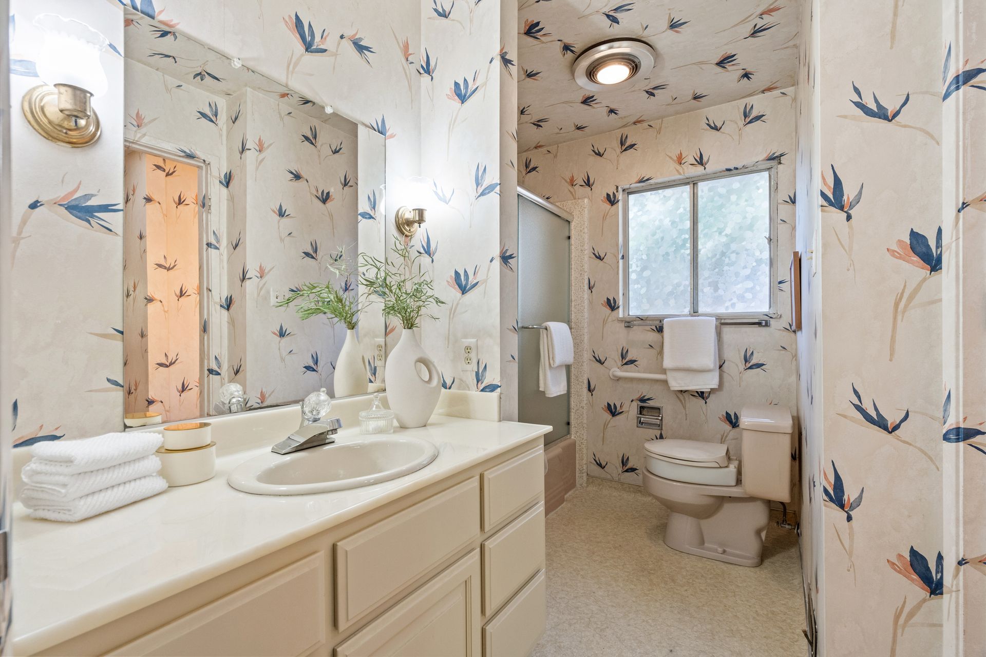 Bathroom with floral wallpaper featuring a sink, toilet, shower, and window; soft white and beige tones.