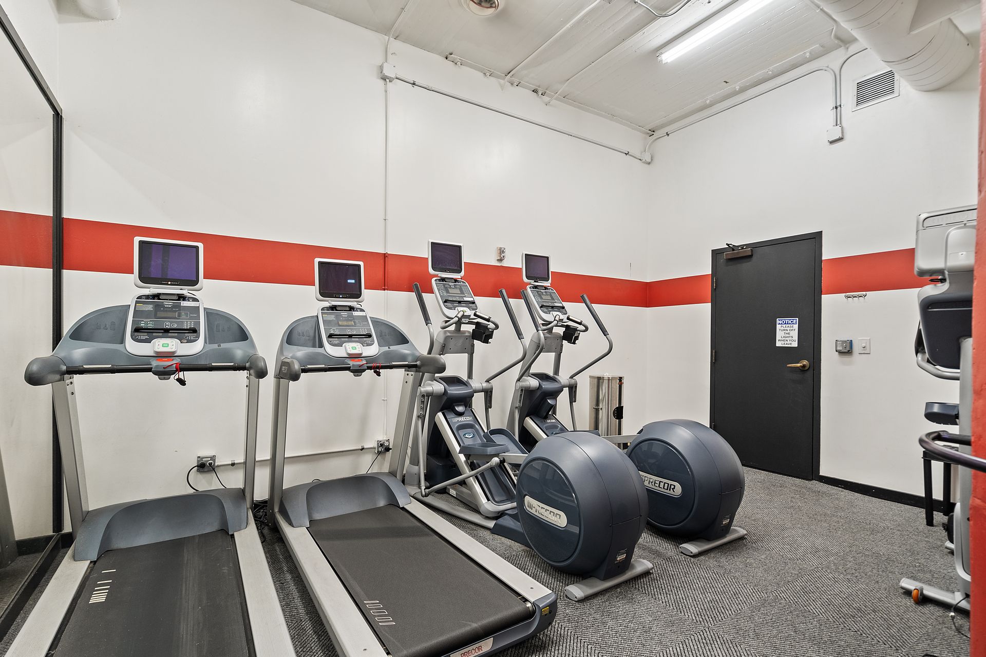 Gym with treadmills, ellipticals, red and white walls, dark door.