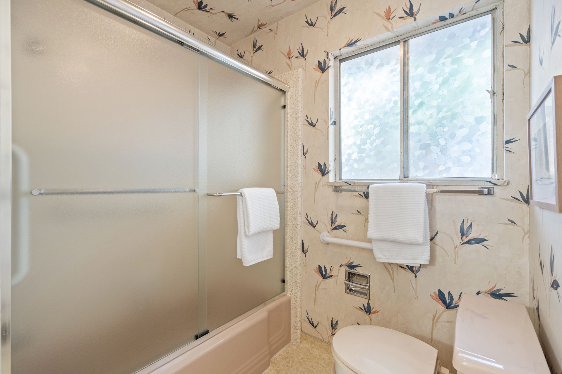 Bathroom with frosted glass shower door, floral wallpaper, and a window above the toilet, with a towel rack.