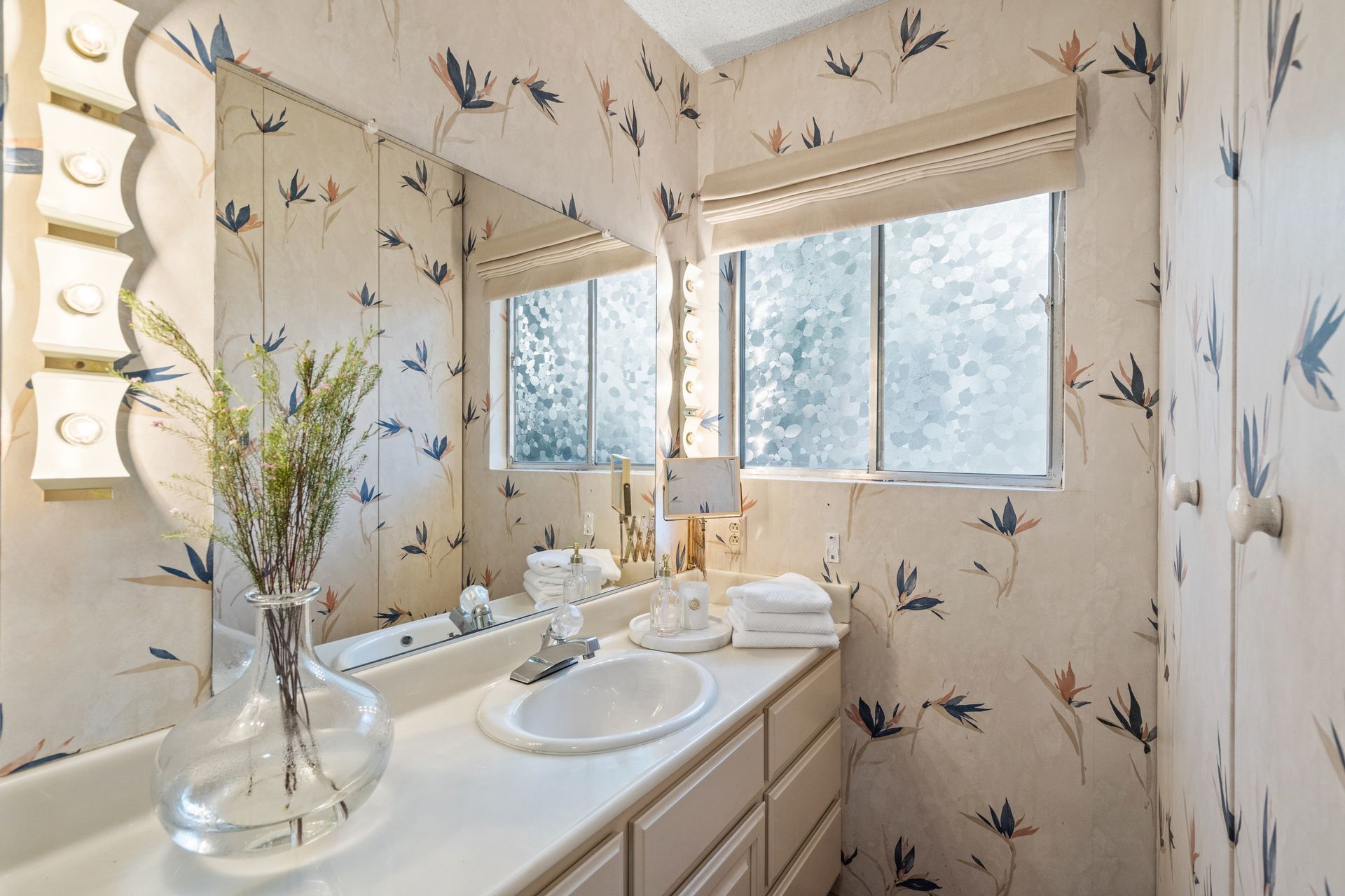 A bathroom with floral wallpaper, a long mirror, a window, and a white countertop with a sink and fresh towels.