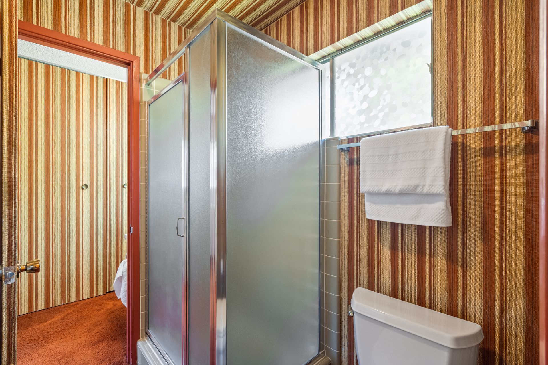 Bathroom with striped brown and orange wallpaper, a glass shower, toilet, and window with a towel.