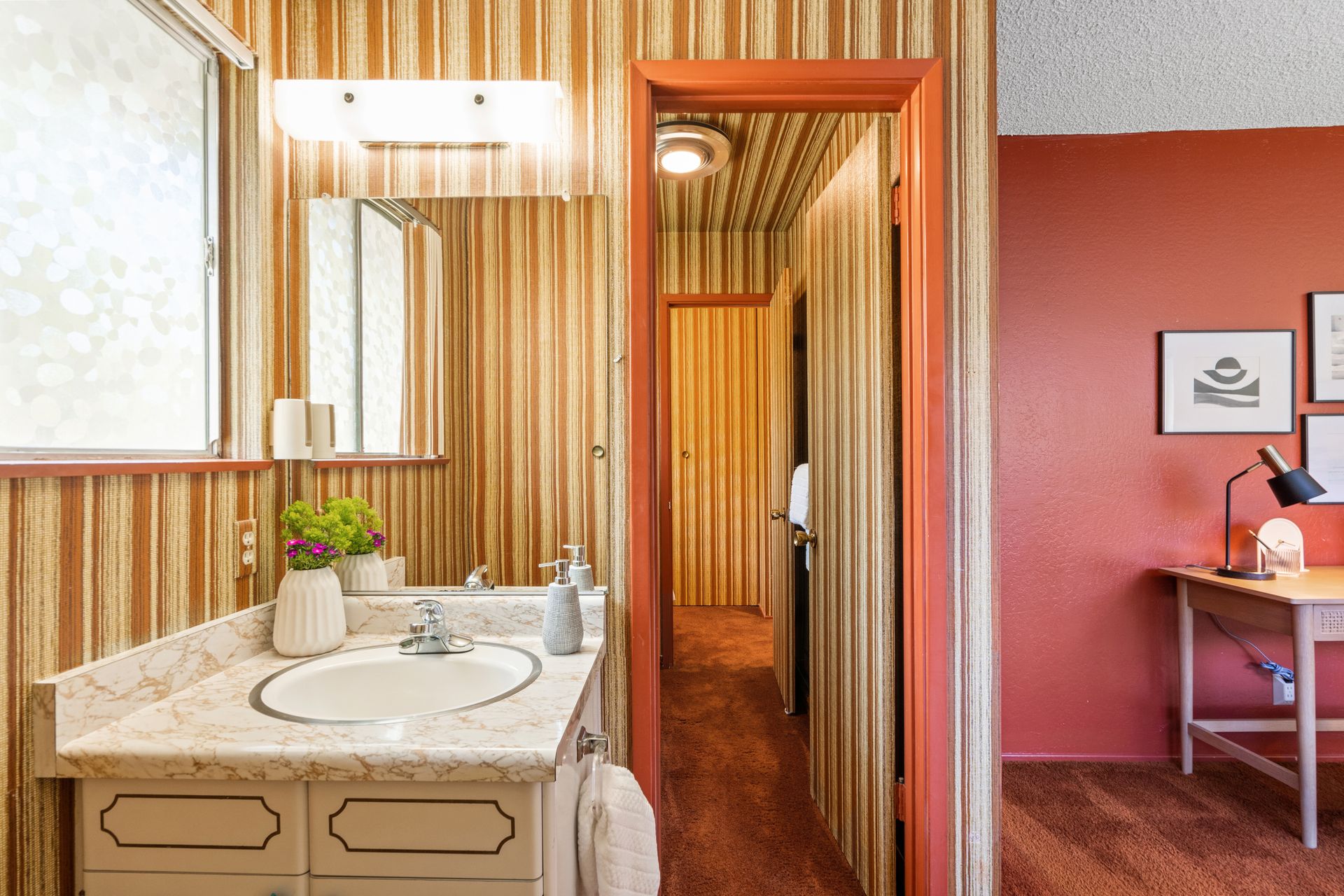 Bathroom with tan and orange striped wallpaper, a vanity, and an open doorway into a hallway with matching wallpaper; to the right is a desk against a red wall.