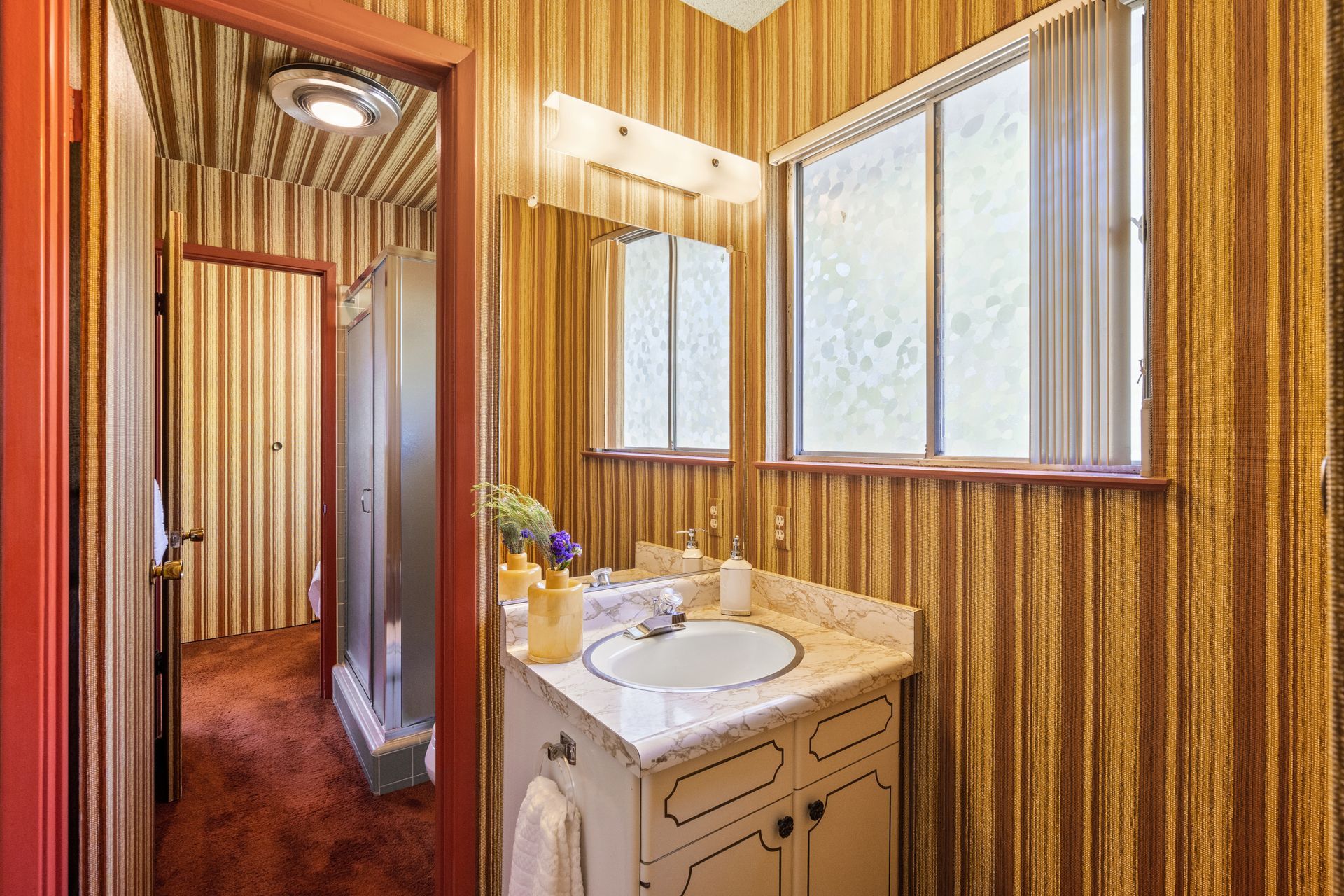 Bathroom with striped wallpaper, a vanity, and a window with frosted glass. A doorway leads to a shower stall.