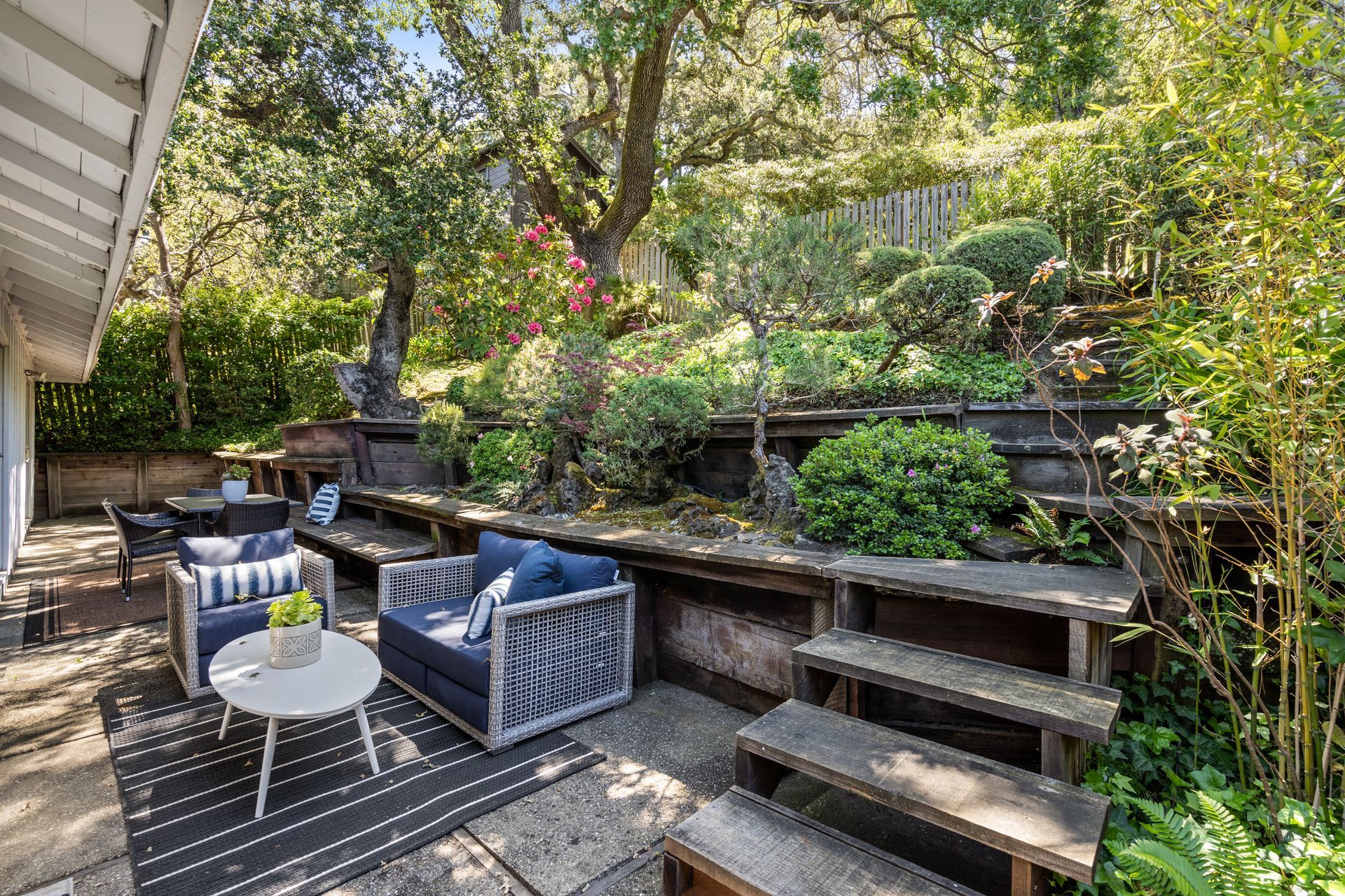 Patio with outdoor furniture, terraced garden with sculpted shrubs, and tall trees on a sunny day.