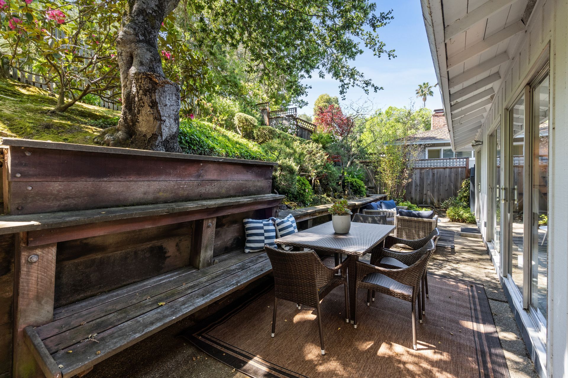 Outdoor patio with a wooden bench, dining table with chairs, and lush greenery.