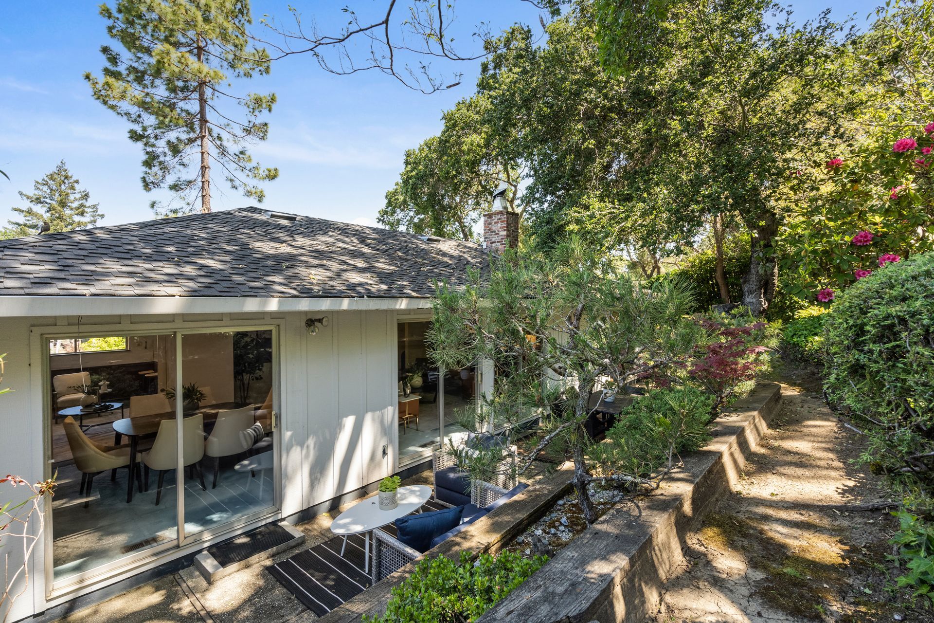 Backyard view of a single-story house with sliding glass doors. A patio area and stairs leading uphill are visible amidst lush greenery.