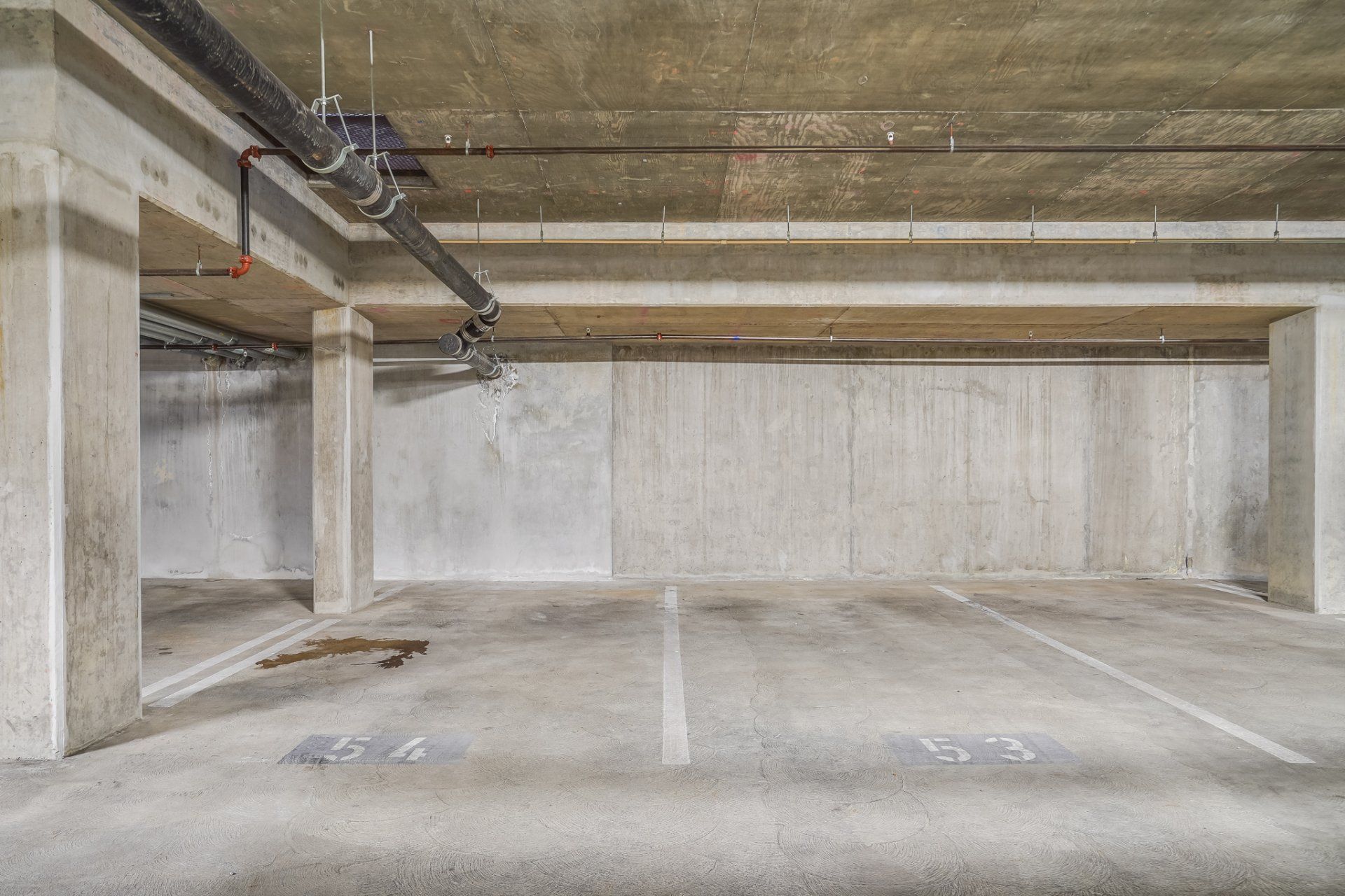 An empty parking garage with concrete walls and pipes hanging from the ceiling.