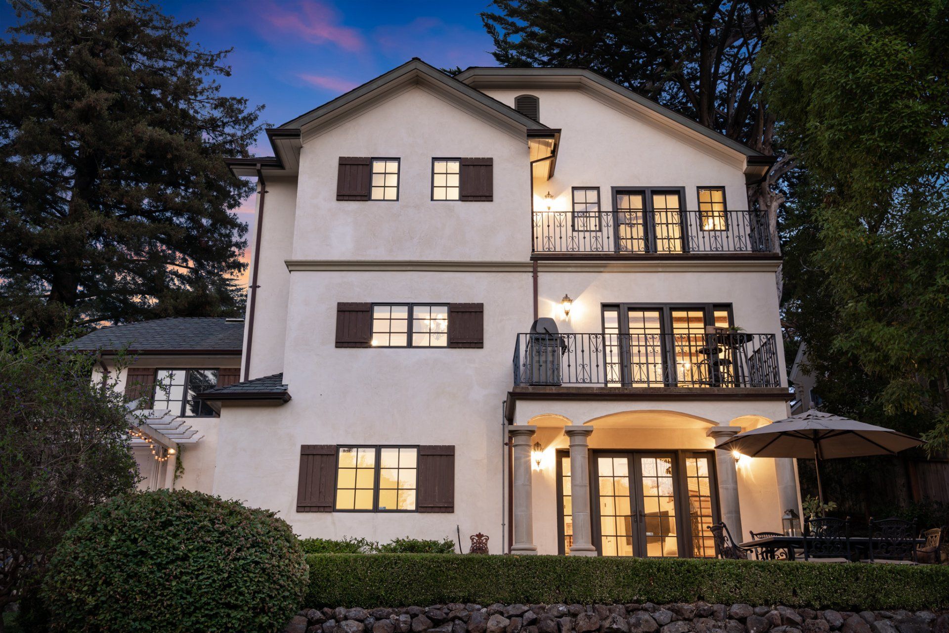 Rear exterior view of a house with brown shutters, balconies, and a patio with an umbrella and table.