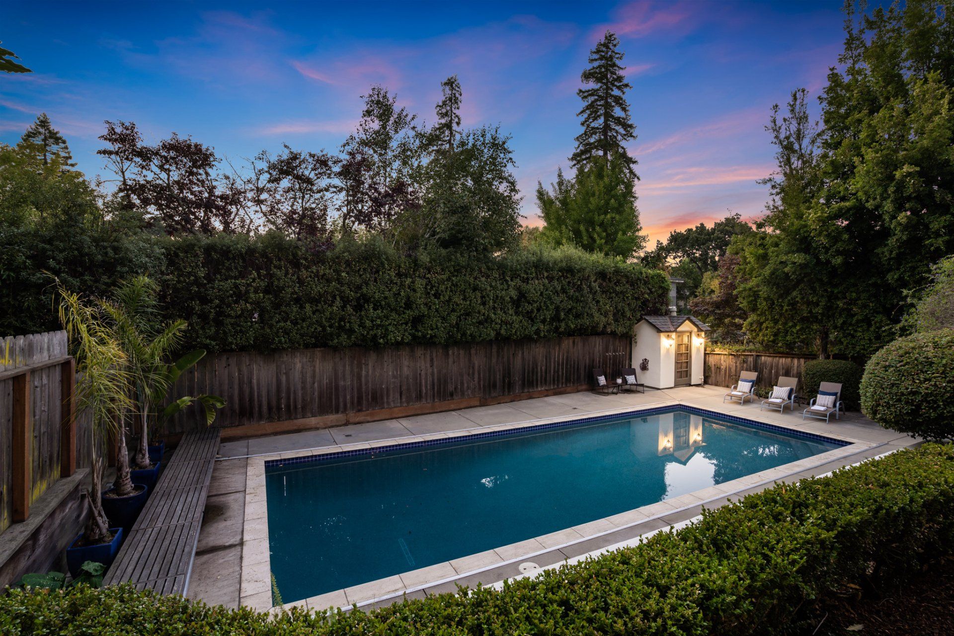 Backyard pool at dusk, surrounded by greenery and a wooden fence. Sky features orange and blue hues.
