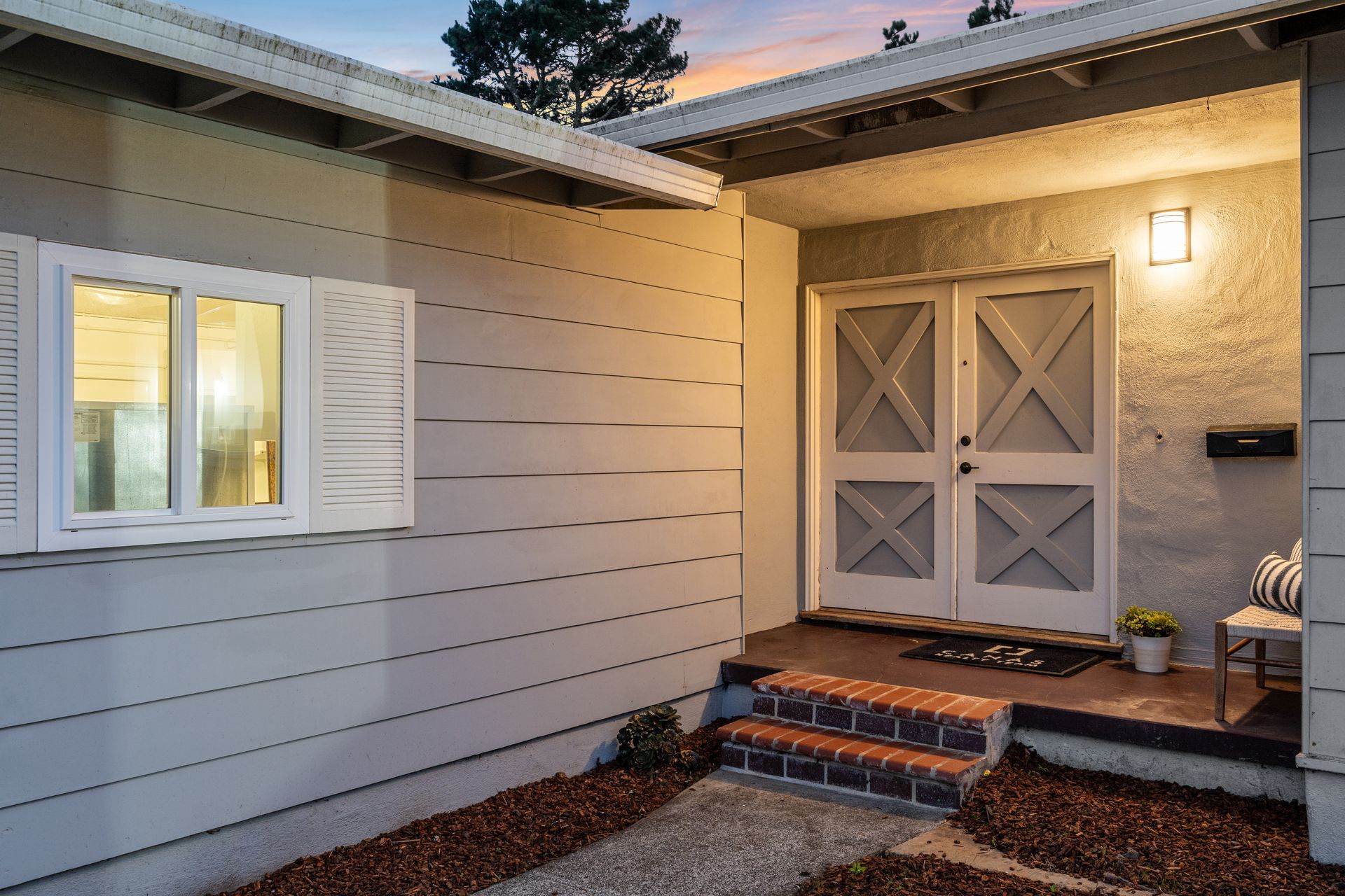 Exterior of a house with white double doors, a porch with steps, and a window with shutters. The walls are light gray.