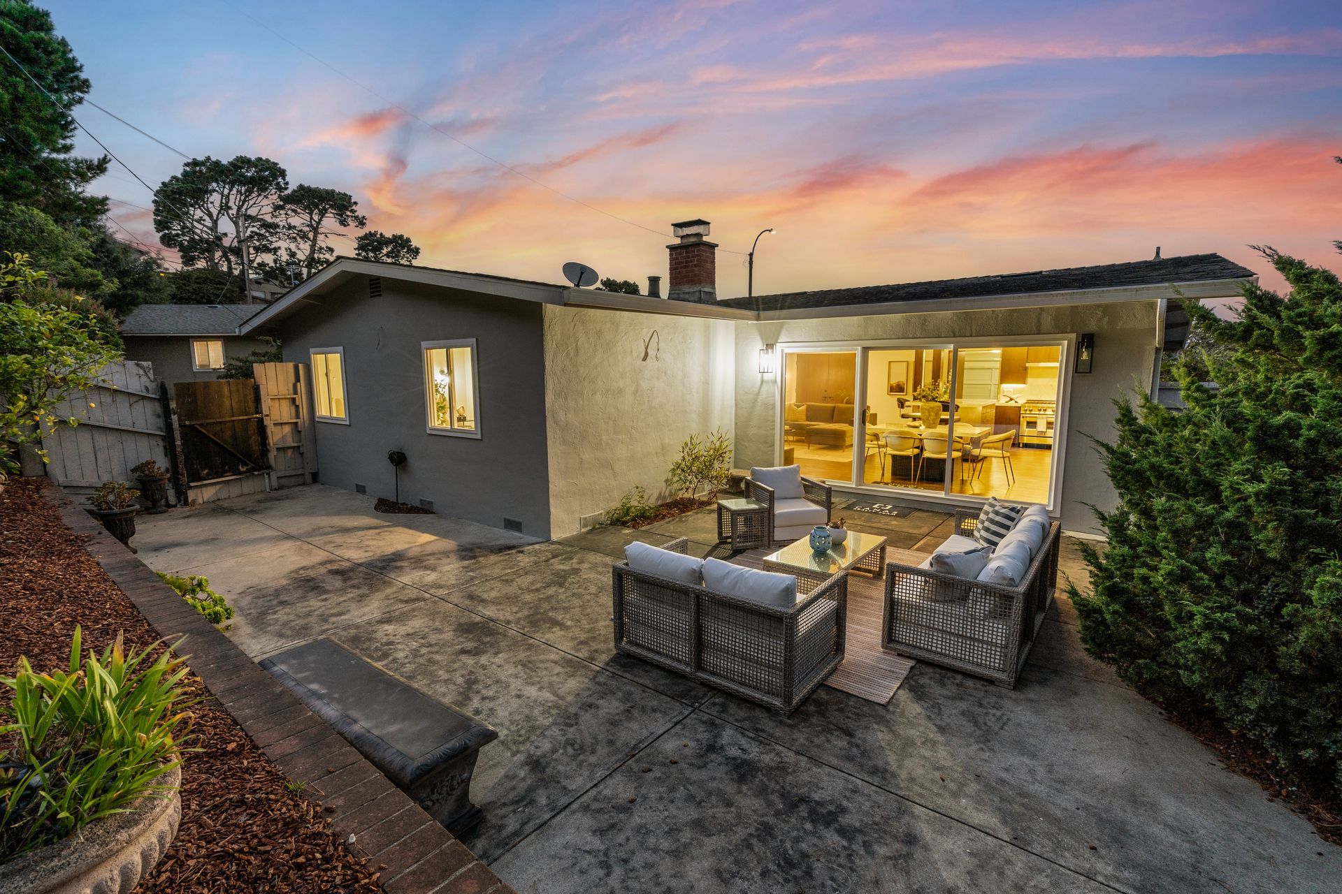 Rear view of a house at dusk with a patio. Grey exterior, lit windows, and outdoor seating under a colorful sky.
