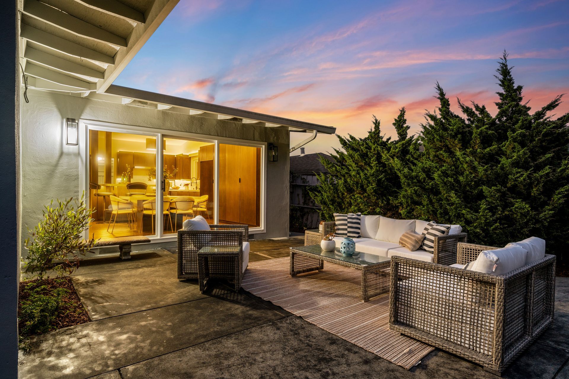 Outdoor patio with seating, rug, and table, adjacent to a house with sliding glass doors. Sunset sky in background.