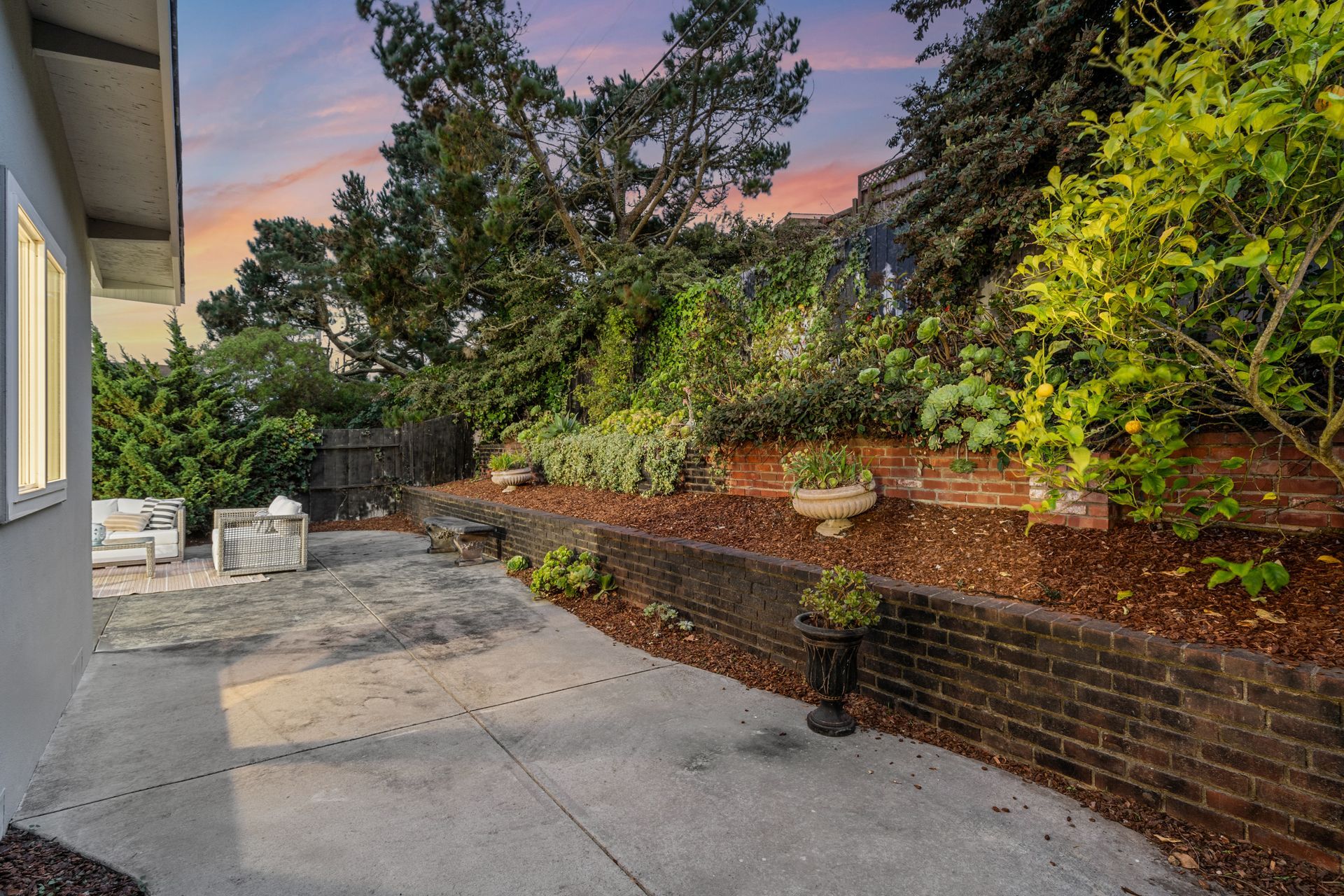 Backyard patio with a brick wall, various plants, and an evening sky.