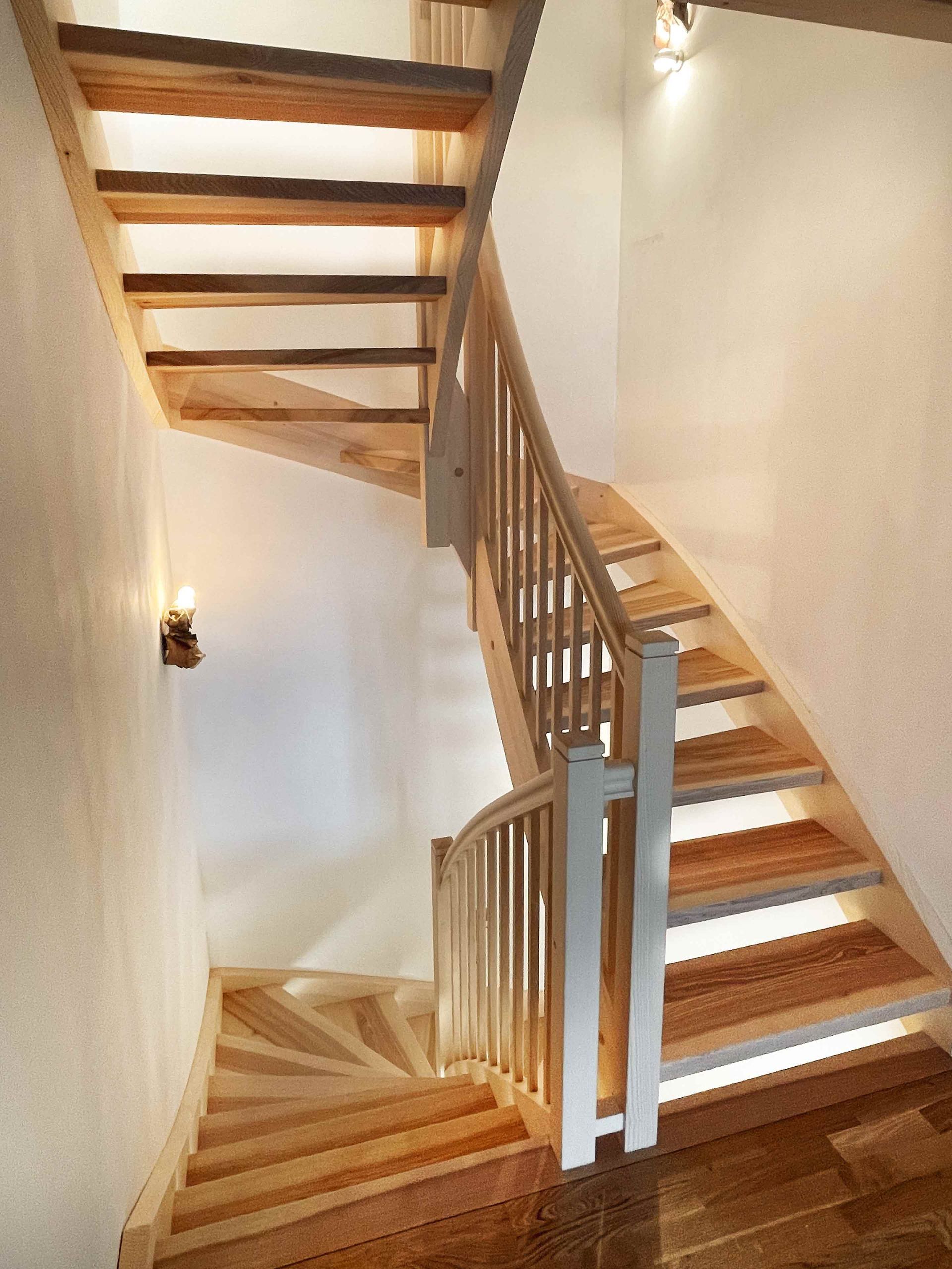 Holztreppe mit weißen Wänden und Geländern, vom unteren Treppenabsatz aus gesehen.