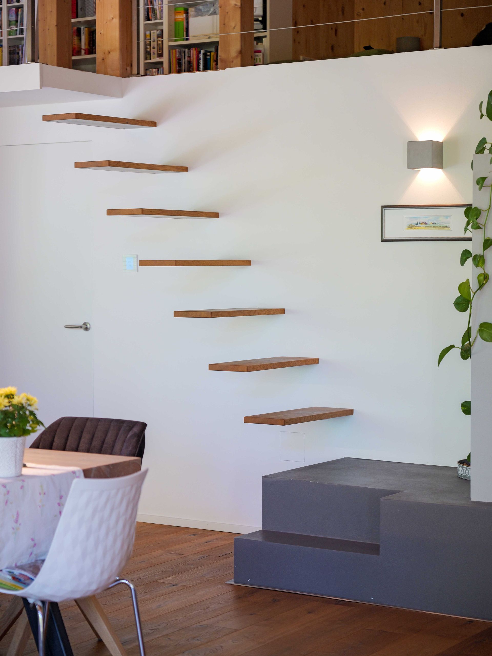 Minimalistisches Interieur mit freitragender Holztreppe, weißer Wand, grauen Sitzmöbeln und einer Wandleuchte.