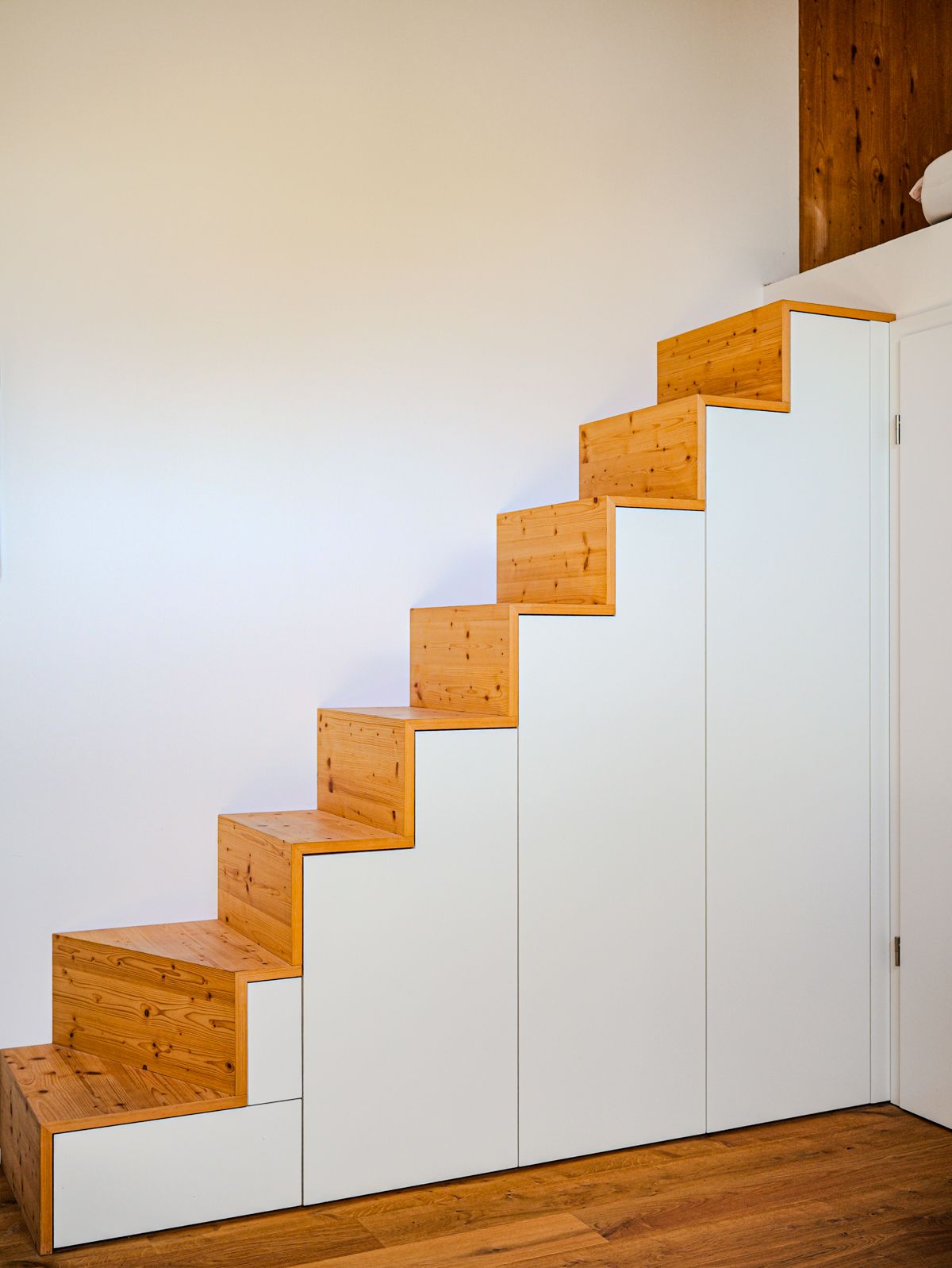 Weiße Treppe mit orangefarbenen Holzstufen und darunterliegenden Einbauschränken, neben einer schlichten Wand.