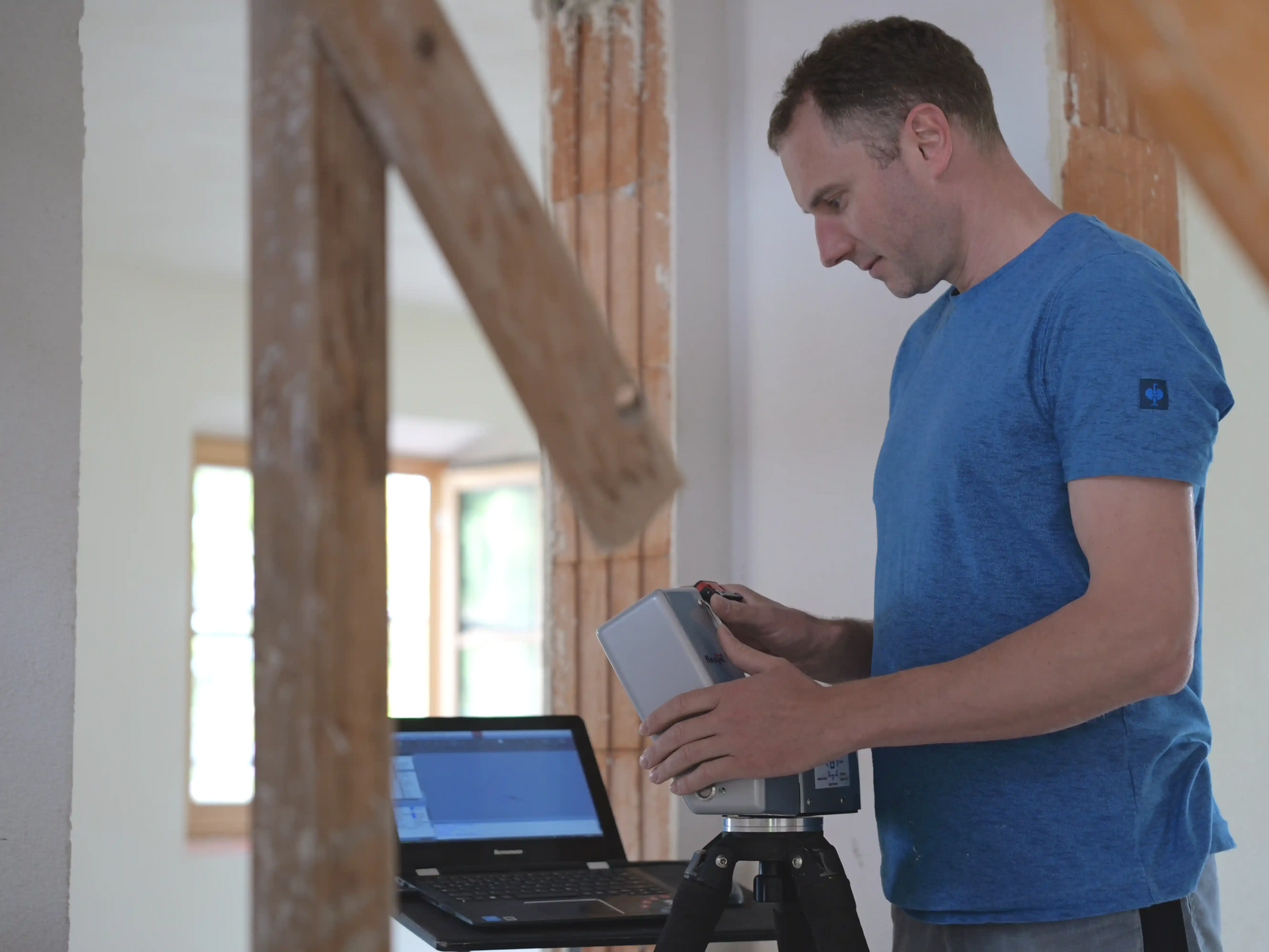 Ein Mann im blauen Hemd justiert ein Gerät auf einem Stativ neben einem Laptop in einem Raum, der gerade renoviert wird.