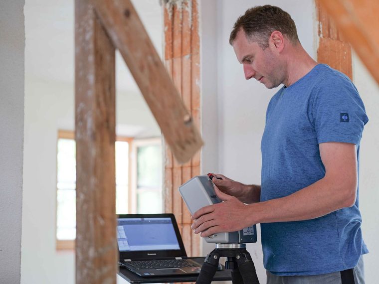 Mann benutzt ein mobiles Gerät neben einem Laptop in einem Raum mit freiliegenden Holzbalken.