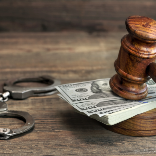 Handcuffs and gavel rest on a stack of hundred-dollar bills on a wooden surface.