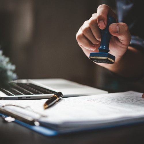 A person stamping a document with a blue ink stamp, next to a laptop and pen.