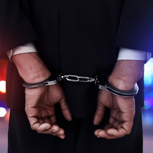 Person in a suit with hands cuffed behind back. Blurred lights in the background suggest a police setting.