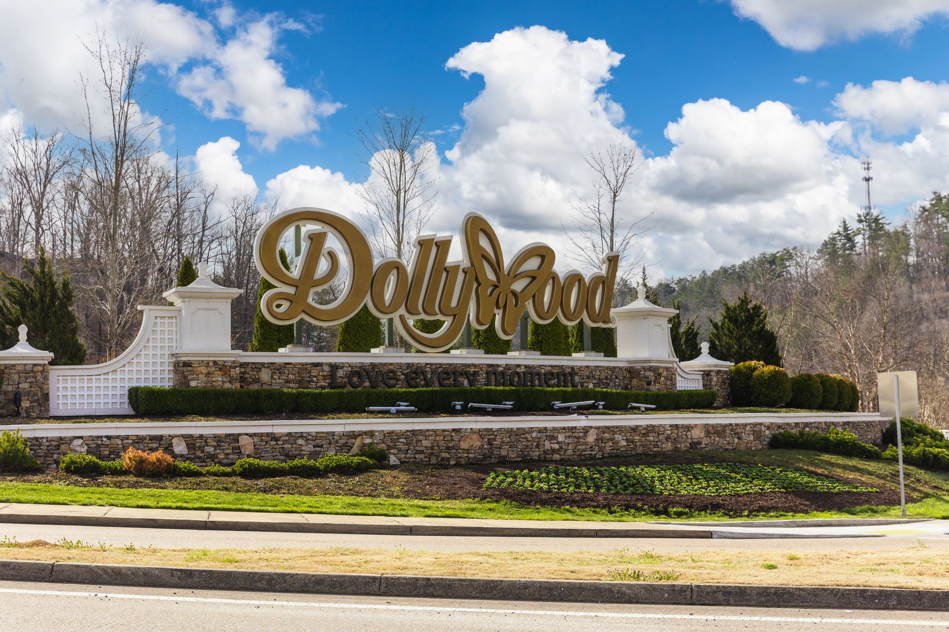 Gold Dollywood logo sign on a stone wall, surrounded by trees and landscaping under a blue sky with white clouds.