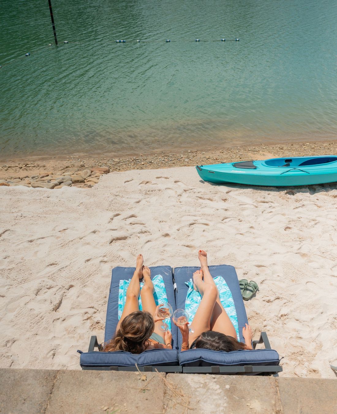 Two people sunbathing on lounge chairs on a sandy beach next to a calm lake with a bright blue kayak nearby.
