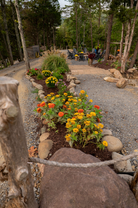 A garden bed with yellow and orange flowers lines a gravel path leading toward a patio with outdoor chairs in the woods.