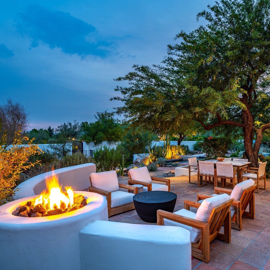 Outdoor patio with fire pit, seating, dining area, and tree. Evening light.