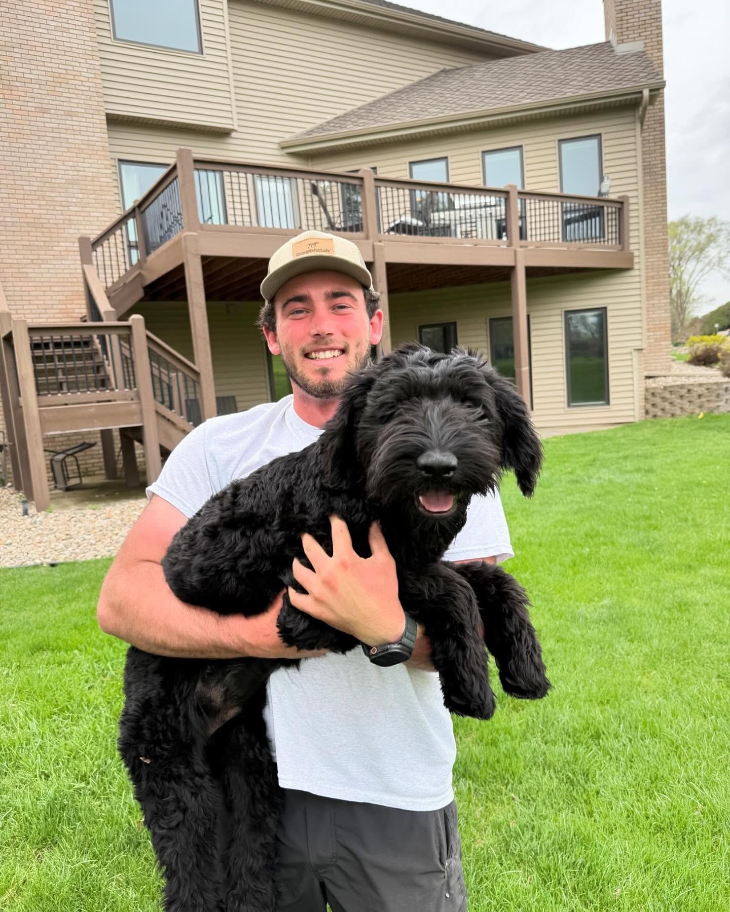 Man smiles holding a black fluffy dog in front of a house with green grass.