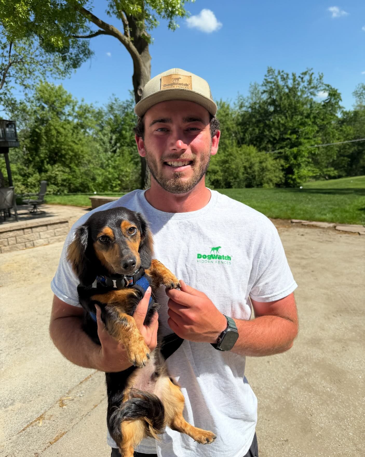 Man holding a dachshund dog outside on a sunny day. Both are smiling.