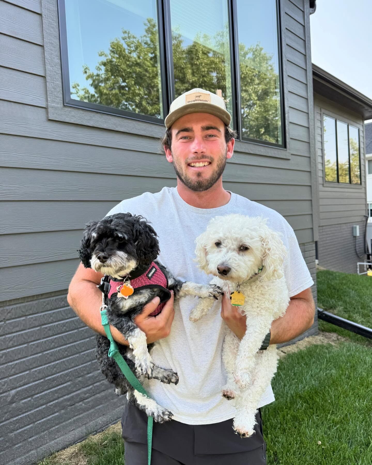 Man holding two small dogs outside a house; one black, one white. He wears a hat and smiles.
