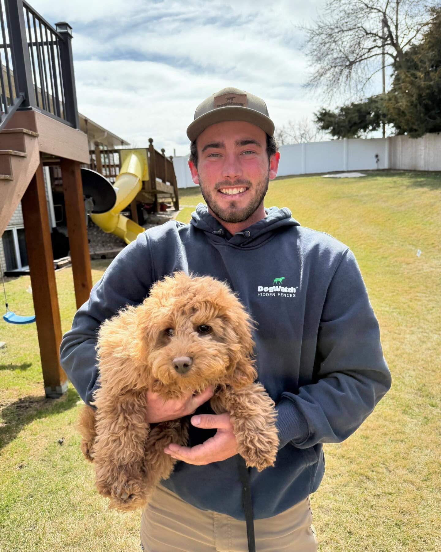 Man holding a small, brown Goldendoodle puppy outside in front of a yard and playset.
