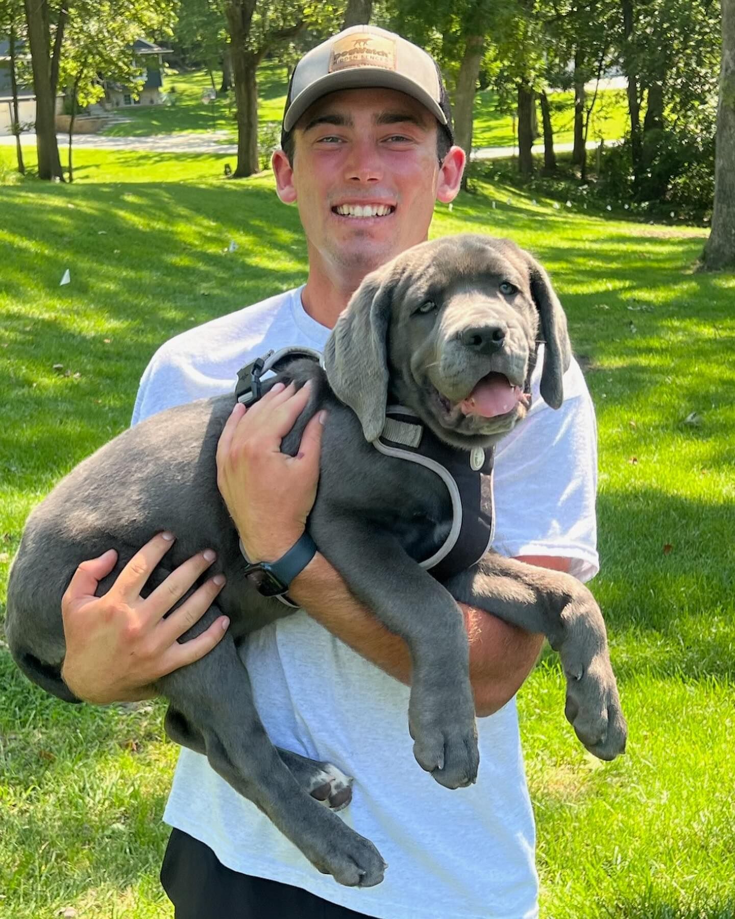 Man holding a gray Great Dane puppy, both smiling, in a grassy outdoor setting.