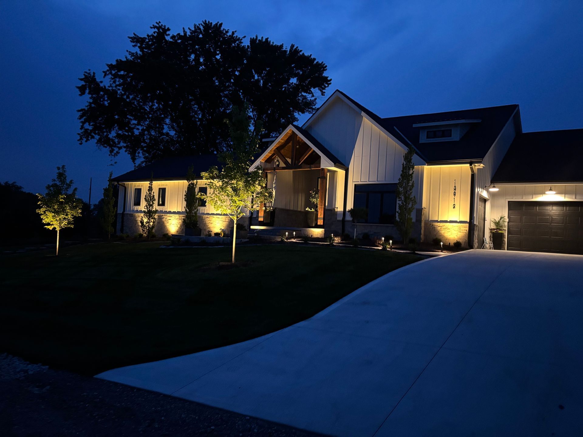 Night view of a modern home with outdoor lighting illuminating the facade and trees. A driveway curves toward the garage.