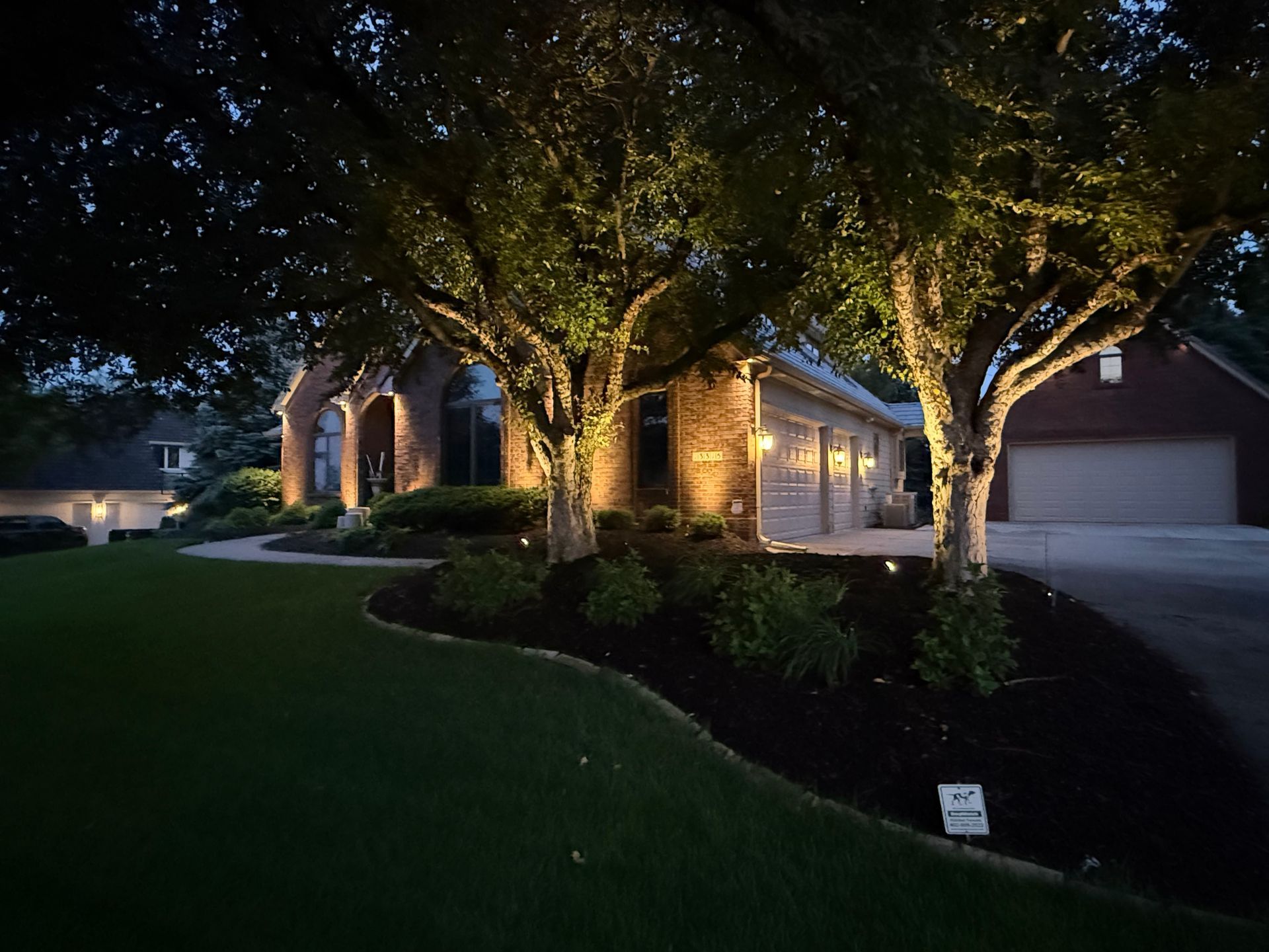 A house at night, illuminated by spotlights on trees, with a green lawn in front.