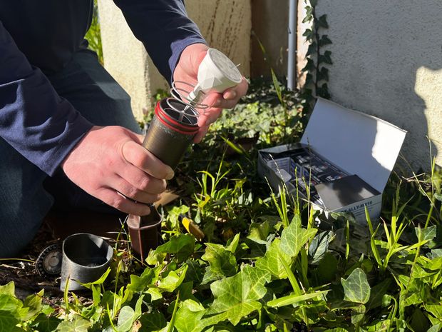 Person assembling a pest control device outdoors near a building with foliage.