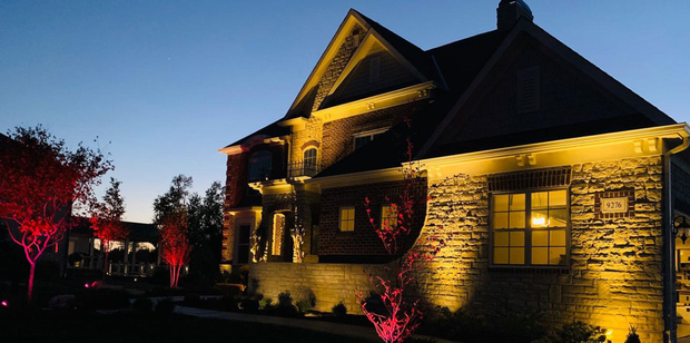 A house is lit up with warm lights against a dusk sky, with red lights illuminating the trees.