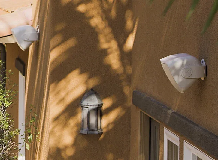 Two white speakers mounted on a stucco wall with shadows. A lantern is also mounted on the wall.