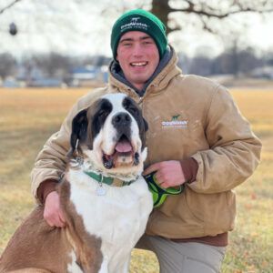 Man in green hat and tan jacket smiles with a Saint Bernard dog in a park.