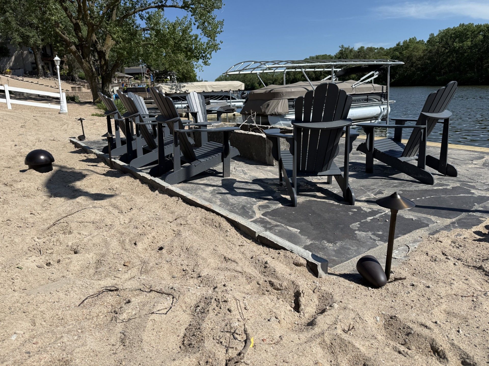 A waterfront seating area with gray Adirondack chairs around a fire pit on a sandy beach.
