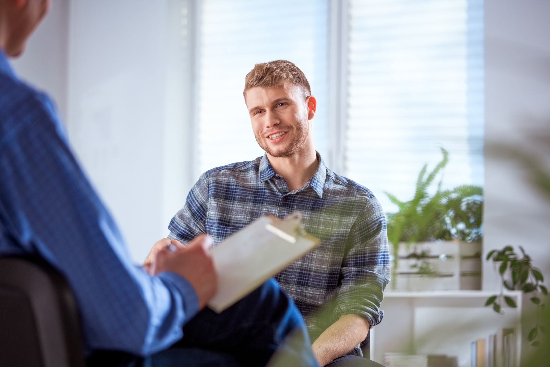 A man is sitting in a chair talking to another man while holding a clipboard.