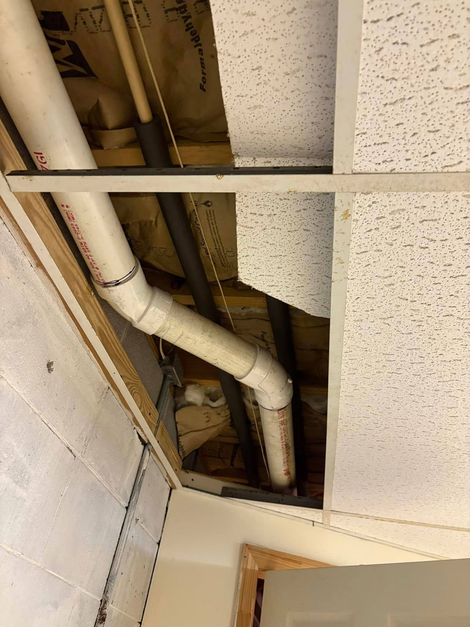 White PVC pipes snaking through an unfinished ceiling with a missing section of ceiling tile and exposed wooden beams.