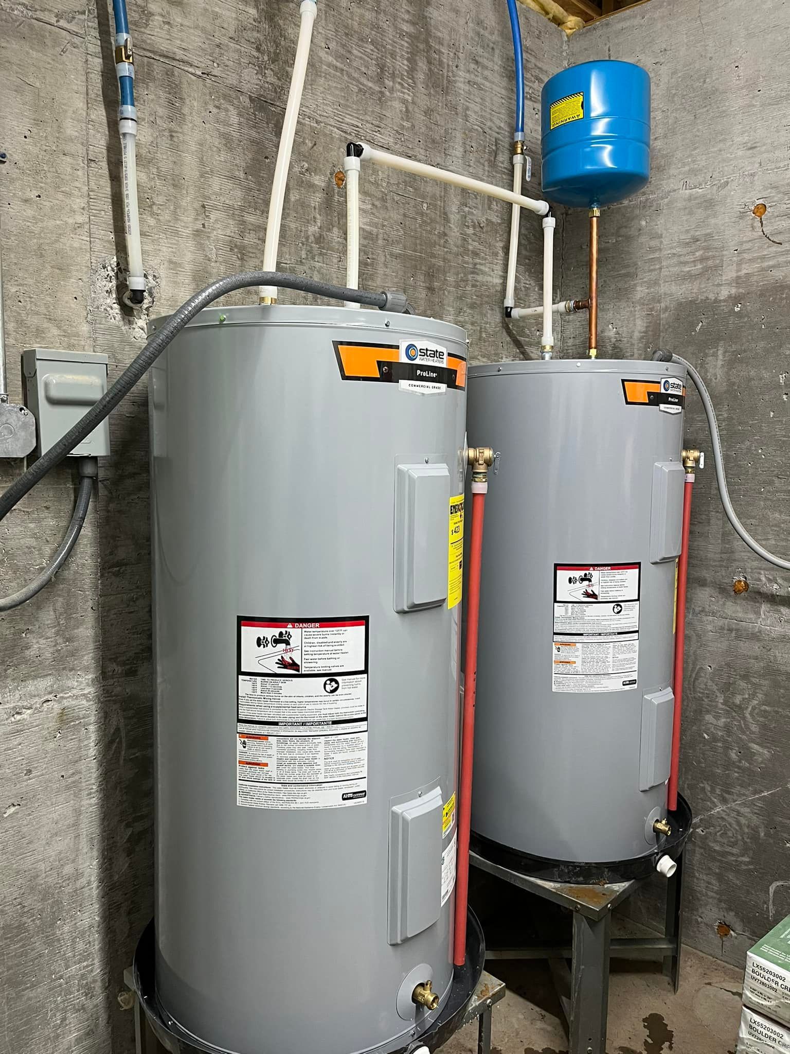 Two gray water heaters stand side-by-side in a concrete room with connected plumbing, a pressure tank, and wiring.