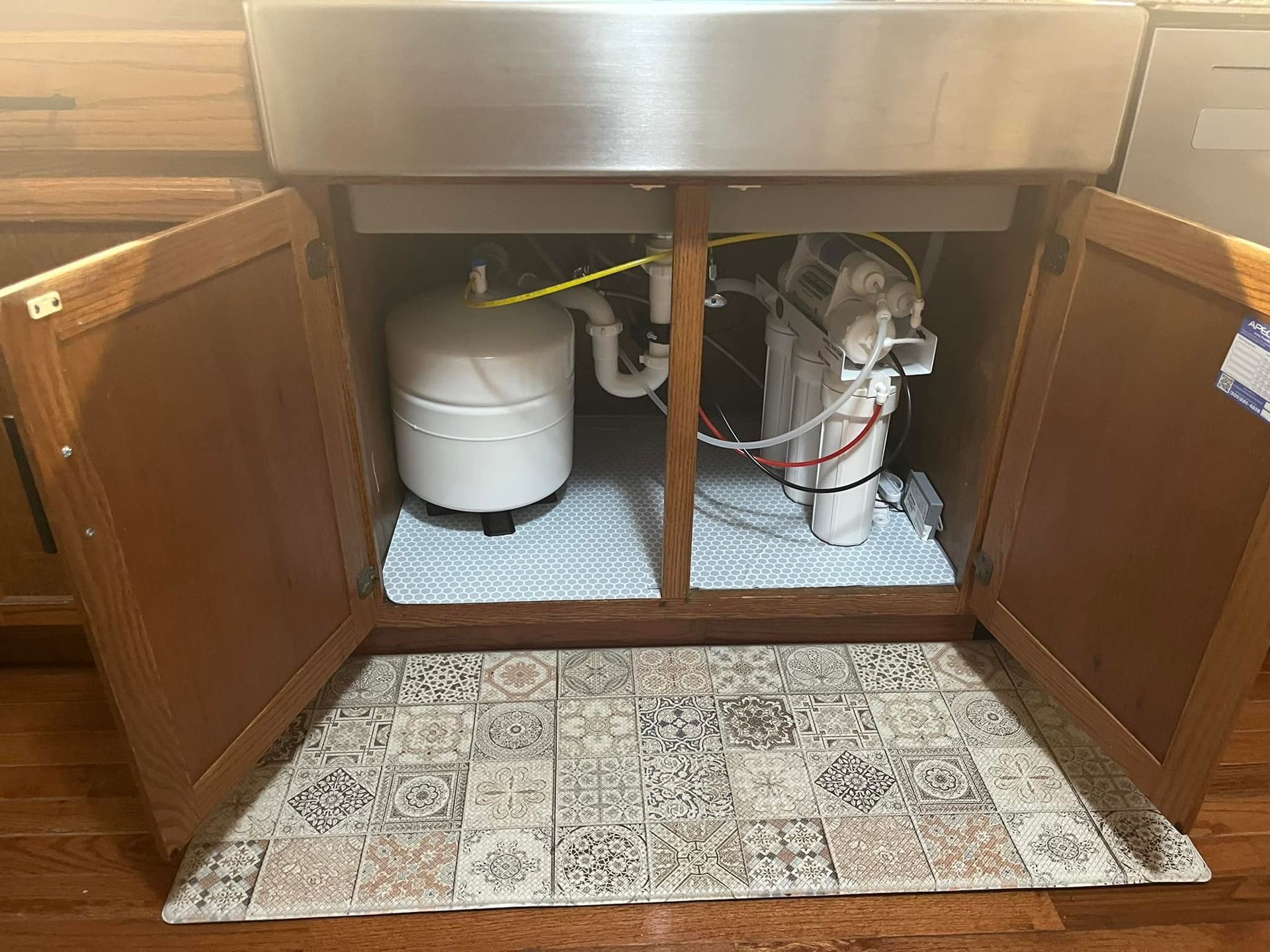 Under-sink view: A stainless steel kitchen sink with cabinet doors open, revealing a water filter system and a pressure tank.