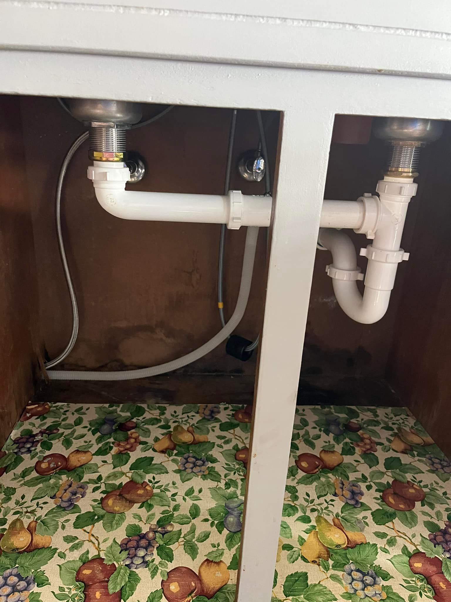 Under-sink plumbing with white pipes, connected to two stainless steel sinks. The cabinet has a patterned floral shelf liner.
