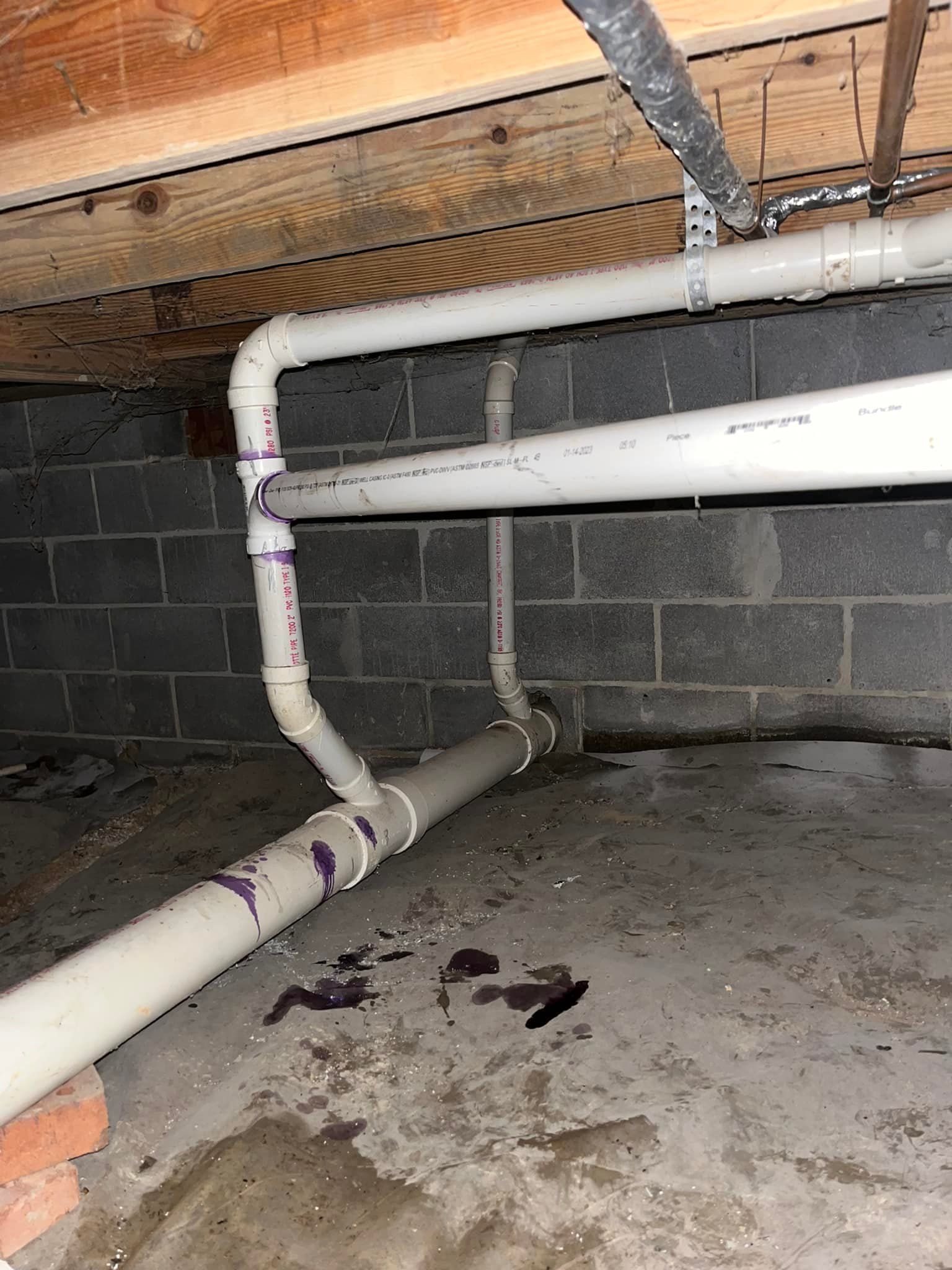 PVC pipes in a crawl space, connected to brick and concrete, with dark stains on the floor.