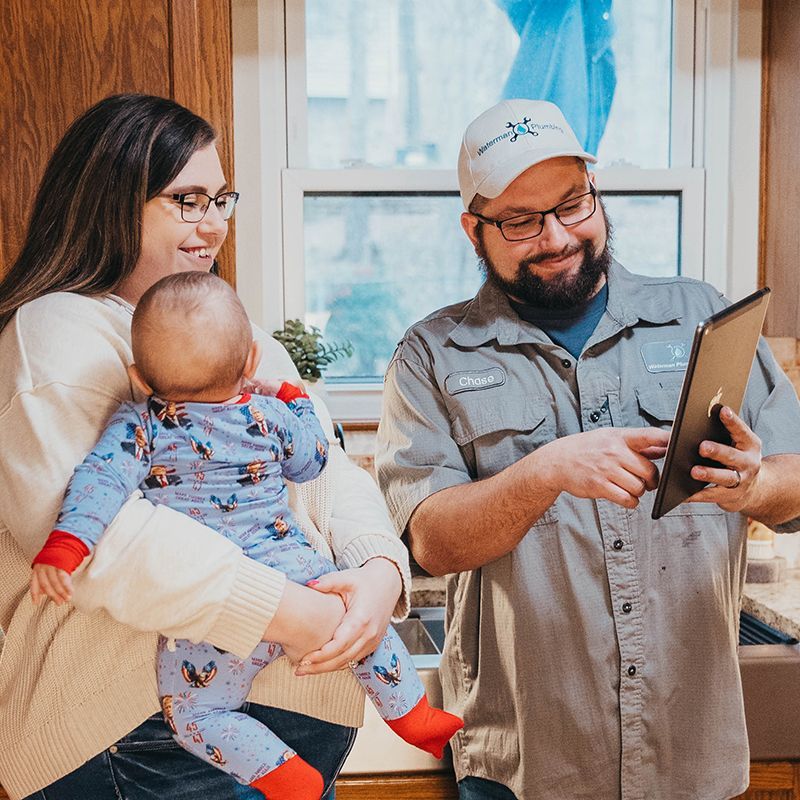 A technician shows a tablet to a woman holding a baby in their kitchen. The technician wears a company hat and uniform.