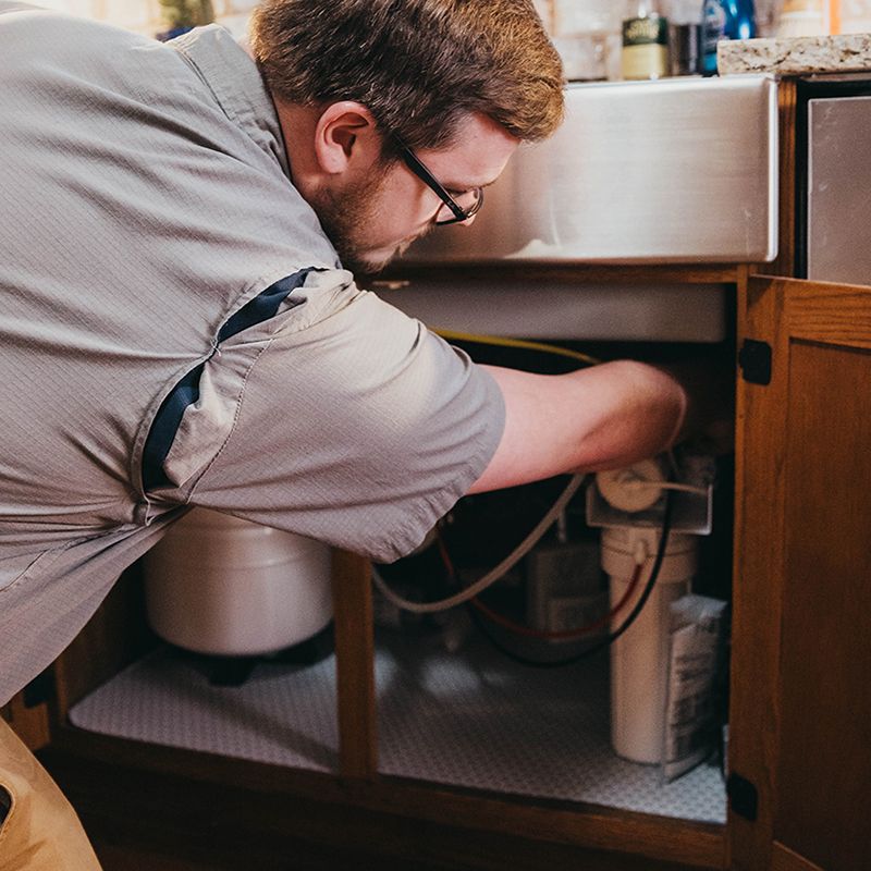 A man with glasses repairs plumbing under a kitchen sink, reaching into the cabinet. He's wearing a gray shirt.