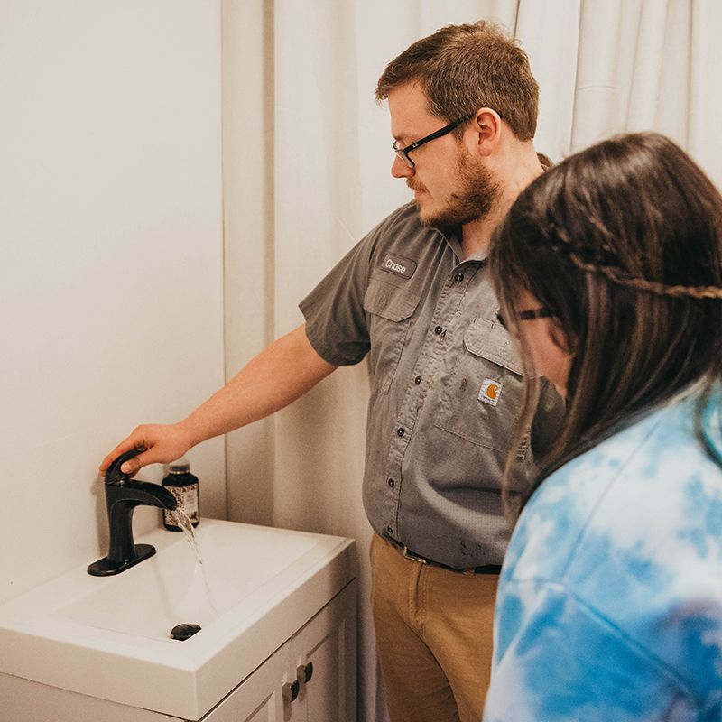 A man in a gray work shirt examines a faucet with a girl looking on. They are in a bathroom with a white sink and curtain.